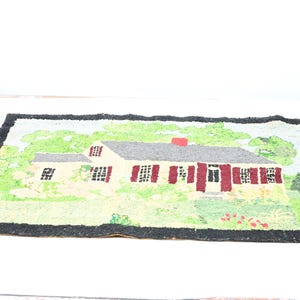 Op de afbeelding: Een rechthoekig tapijt met een gedetailleerde afbeelding van een huis. Het huis heeft een grijs dak, rode luiken en een rode schoorsteen. Het tapijt heeft een zwarte rand en een groen en gele achtergrond.