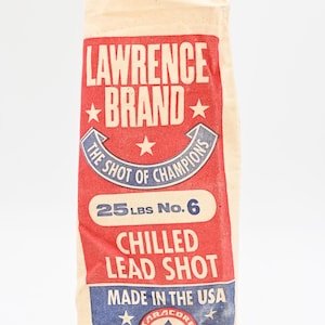 May include: A white canvas bag with red, blue, and white text and stars. The bag is labeled "Lawrence Brand", "The Shot of Champions", "25 lbs No. 6", "Chilled Lead Shot", "Made in the USA", and "Taracorp Industry".