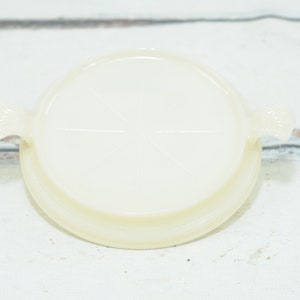 Puede incluir: Un plato para tartas de vidrio blanco con dos asas. El plato tiene un centro elevado y un borde ligeramente festoneado.