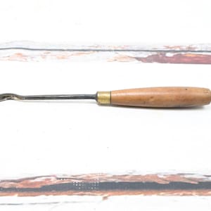 May include: A vintage woodworking tool with a curved metal blade and a wooden handle. The handle is light brown and cylindrical, with a brass ferrule where it meets the metal shaft. The tool is designed for carving or shaping wood.