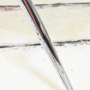 May include: Close-up of a chrome metal chair leg, angled diagonally across the frame. The leg has a shiny, reflective surface with minor wear and tear. The background is a white, weathered wooden surface.