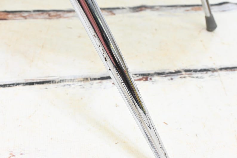 May include: Close-up of a chrome metal chair leg, angled diagonally across the frame. The leg has a shiny, reflective surface with minor wear and tear. The background is a white, weathered wooden surface.