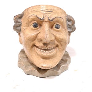 May include: A beige ceramic head planter featuring a smiling face with wide eyes, arched eyebrows, and a ruffled collar base. The facial features are painted with dark brown accents, and the surface has a textured, weathered appearance.