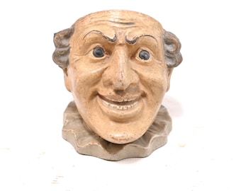 May include: A beige ceramic head planter featuring a smiling face with wide eyes, arched eyebrows, and a ruffled collar base. The facial features are painted with dark brown accents, and the surface has a textured, weathered appearance.