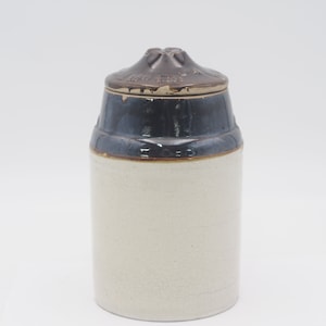 May include: A vintage stoneware jar with a lid. The jar has a cylindrical shape with a light beige body and a dark blue band around the top. The lid is brown with a decorative design. The jar is likely used for storage.