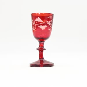 May include: A ruby red wine glass with a stem and a round base. The glass features an etched design of grapes and leaves. The glass is made of a transparent material and is set against a white background.