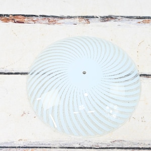 May include: A light blue glass lampshade with a spiral design. The shade has a central hole and is set against a white wooden background. The spiral pattern is made of white lines.