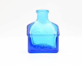 Peut inclure: Une petite bouteille carrée en verre bleu avec un dessus bleu clair. La bouteille a une surface texturée et les mots "Co. 1891" sont visibles sur le fond.