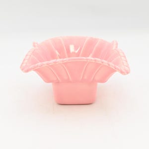 May include: A light pink ceramic dish with a scalloped rim and a square base. The dish has a fluted design on the inside and a textured edge. The overall shape is reminiscent of a flower.