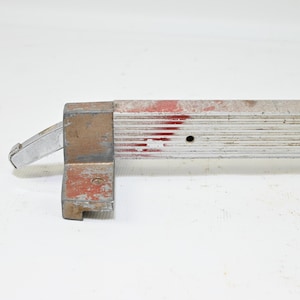 Peut inclure: Une barre rectangulaire métallique à texture nervurée, présentant une finition rouge et argentée usée. Une solide équerre en forme de L avec une patine gris-brun vieillie et un petit levier métallique argenté saillant est fixée à une extrémité.
