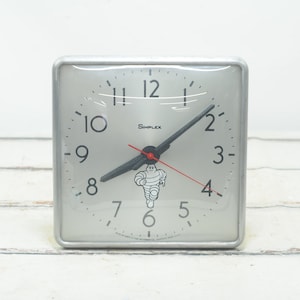 May include: A silver square clock with black hands and a red second hand. The clock face has black numbers and the word "Simplex" printed on it. A black and white illustration of the Michelin Man is in the center of the clock face.