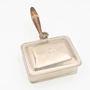 Op de afbeelding: Een zilverkleurige rechthoekige asbak met een houten handvat. De asbak heeft een decoratieve rand en een wapenontwerp op het deksel. De handgreep is lichtbruin.
