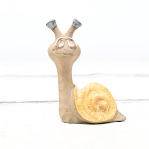 May include: A ceramic snail figurine with a yellow shell and brown body. The snail is wearing glasses and has a smiling face.