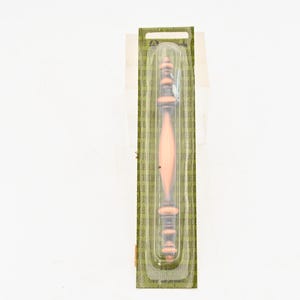 May include: A wooden spindle-shaped object with a light brown center and darker brown ends, encased in clear plastic. The object is packaged on a green and tan patterned backing.