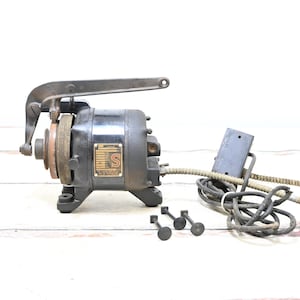May include: An antique black electric motor with a handle and a pulley. The motor has a metal plate with text and is connected to a power cord and a control box. Three small metal pieces are in front of the motor.