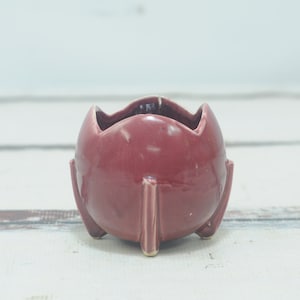 Pode incluir: Um pequeno vaso de cerâmica vermelho com formato arredondado e quatro pernas. O vaso tem um acabamento liso e brilhante.
