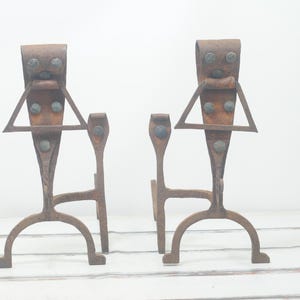 Puede incluir: Un par de soportes de chimenea de metal rústico con un diseño triangular y correas de cuero. Los soportes están hechos de hierro y tienen un acabado marrón oscuro.
