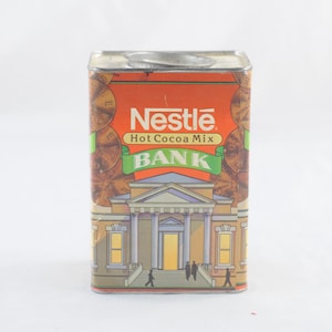 Pode incluir: Um cofrinho de lata vermelho, branco e verde com uma imagem de um pr&eacute;dio e as palavras "Nestle Hot Cocoa Mix Bank" na frente.