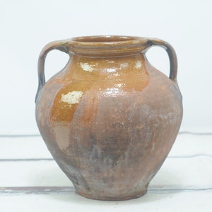 May include: A brown ceramic pitcher with two handles. The pitcher has a rustic, aged look and is glazed in a light brown colour.