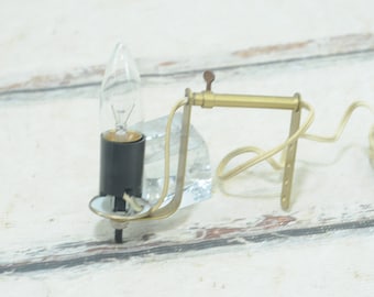 May include: A gold-toned wall sconce with a clear glass shade and a clear light bulb. The sconce has a simple design and is attached to a white wall.