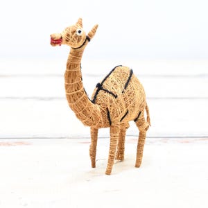 May include: A small, hand-crafted camel figurine made from natural materials. The camel is brown and has a black saddle. It is standing on a white surface.