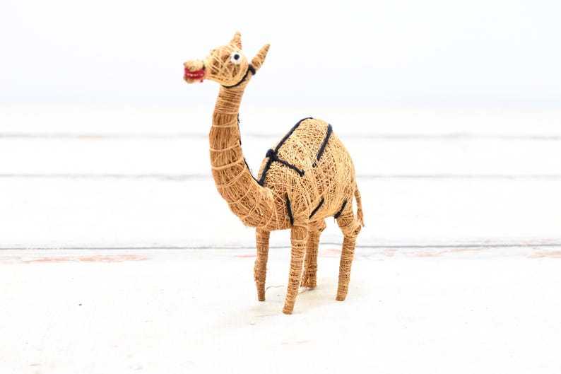 May include: A small, hand-crafted camel figurine made from natural materials. The camel is brown and has a black saddle. It is standing on a white surface.
