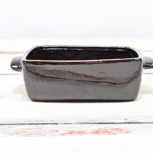 May include: A rectangular, dark brown ceramic dish with rounded handles on each side. The dish has a glossy finish and is likely used for baking or serving food. The interior is a darker shade of brown.