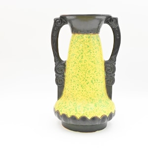 May include: A decorative vase with a speckled yellow body and black accents. The vase has two ornate handles and a scalloped base. The top and base are black, while the body is yellow with green speckles. A unique piece for home decor.