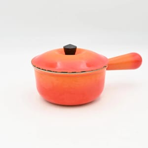 Pode incluir: Uma pequena panela de ferro fundido esmaltado laranja com tampa e alça. A panela tem formato arredondado e um botão preto na tampa. A alça é curva e se estende da lateral. Fundo branco.