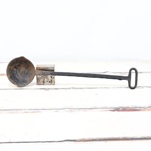May include: An antique, cast iron ladle with a round bowl and a long, straight handle ending in a loop. The ladle has a dark, aged appearance, suggesting it is vintage. The handle is attached to the bowl with a rectangular piece.
