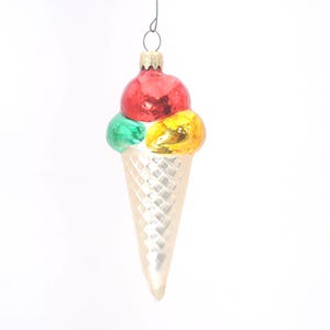 May include: A glass ice cream cone ornament featuring three scoops in red, green, and yellow. The cone has a textured, diamond-patterned surface in a pale cream color. A gold-toned metal cap sits at the top with a wire loop for hanging.