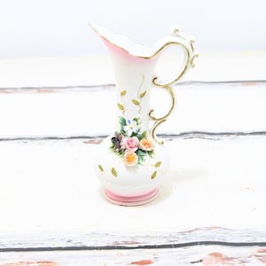 May include: A small white porcelain jug with a pink rim and gold accents. The jug features a floral design with pink, yellow, and white flowers and green leaves.