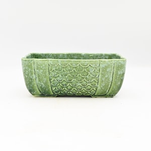 May include: A rectangular, green ceramic planter with a textured, scaly pattern on the front. The planter has a slightly weathered appearance and is suitable for indoor or outdoor use. The planter is approximately 10 inches long.