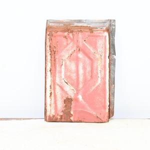 May include: A weathered, rectangular metal piece with a faded pink front and a geometric design. The edges are a contrasting gray, showing signs of rust and age, suggesting a vintage or antique item.