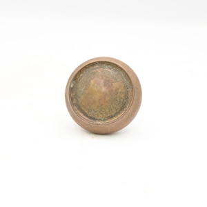 Könnte beinhalten: Ein runder, antiker Schrankknauf aus Bronze. Der Knauf hat eine kreisförmige Form mit einer leicht gewölbten Oberseite und einem flachen Boden. Die Oberfläche weist Alters- und Abnutzungserscheinungen auf, mit einem fleckigen, gealterten Aussehen.