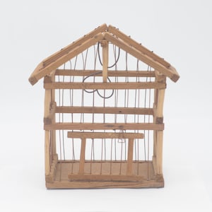 Puede incluir: Una pequeña jaula de pájaros de madera con una malla de alambre en el interior y una percha de madera. La jaula tiene un techo a dos aguas y está hecha de madera natural.