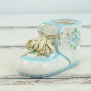 Könnte beinhalten: Ein weißer Keramik-Babyschuh-Pflanztopf mit blauen Verzierungen und floralen Details. Der Schuh hat ein gelbes Band gebunden.