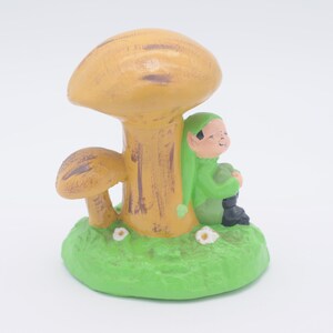 Può includere: Una statuetta in ceramica di un gnomo seduto sotto un fungo. Lo gnomo indossa un cappello verde e abiti verdi. Il fungo è giallo e marrone.