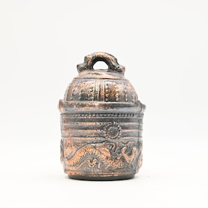 Puede incluir: Una campana decorativa de color bronce con un asa en la parte superior. La campana presenta tallas en relieve detalladas de dragones y otros diseños ornamentados. La campana tiene una apariencia desgastada, lo que sugiere edad o un estilo vintage.