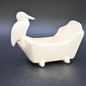 Puede incluir: Una maceta de cerámica color crema con forma de bañera, con un pájaro posado en la parte trasera. La maceta tiene forma rectangular con bordes redondeados y se apoya en cuatro patas pequeñas. El pájaro tiene un pico largo y mira hacia la izquierda.