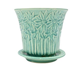 Puede incluir: Una maceta de cerámica de color turquesa claro con su plato a juego. El exterior de la maceta presenta un patrón floral en relieve de margaritas alrededor del borde superior, que se transforma en líneas verticales hacia la base.