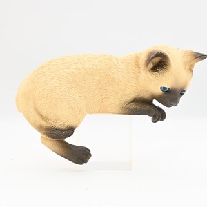 May include: A Siamese cat figurine with a light tan body, dark brown ears, paws, and face, and bright blue eyes. The cat is posed as if it is looking down, with its front paws over the edge of a white surface.