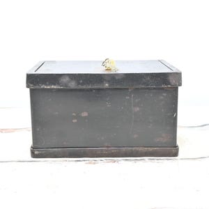 May include: A rectangular, black metal box with a hinged lid. The box has a keyhole and a small key on top. The box is likely vintage or antique, with signs of wear and tear. The box is sitting on a white surface.