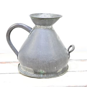 May include: A vintage metal pitcher with a large handle and a small loop handle on the side. The pitcher has a wide base and a narrow neck with a flared rim. The metal has a weathered, aged appearance.