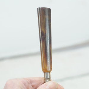 Op de afbeelding: Een bruin en zwart hoornvormig object met een metalen punt. Het object wordt in een hand gehouden.