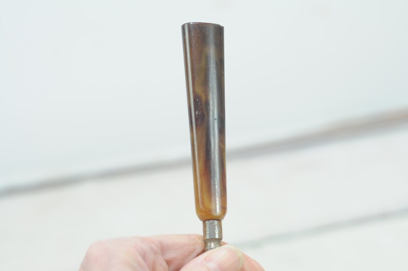 Op de afbeelding: Een bruin en zwart hoornvormig object met een metalen punt. Het object wordt in een hand gehouden.