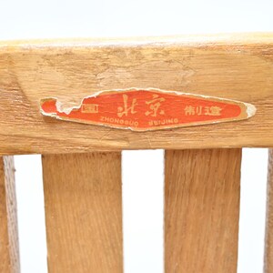May include: Close-up of a wooden chair back with a faded orange sticker. The sticker has Chinese characters and the words "Beijing" and "Zhongguo" in English. The wood grain is visible, and the chair back has vertical slats.