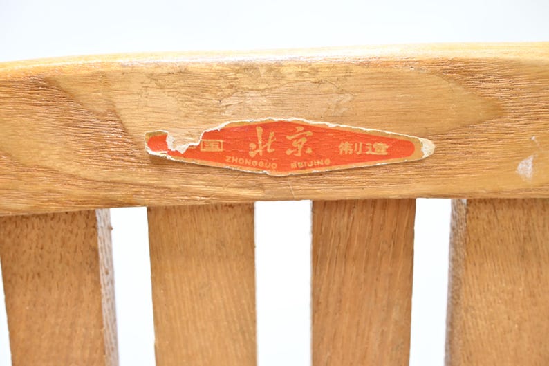 May include: Close-up of a wooden chair back with a faded orange sticker. The sticker has Chinese characters and the words "Beijing" and "Zhongguo" in English. The wood grain is visible, and the chair back has vertical slats.