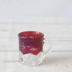 May include: A small, clear glass cup with a red, ruby-colored rim and a handle. The cup has a diamond-shaped pattern and is etched with the words "Lilly a -1902."