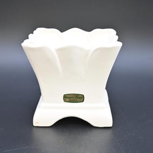 Puede incluir: Un jarrón de cerámica blanco con borde festoneado y base cuadrada. El jarrón tiene una forma geométrica acampanada y una etiqueta en la parte delantera. El jarrón mide aproximadamente 15 cm de alto.